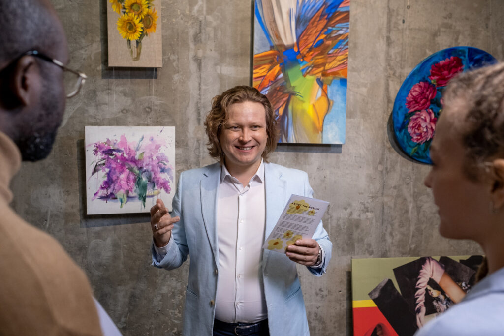 Happy modern artist with leaflet guide presenting collection of his artworks to young intercultural guests of art gallery