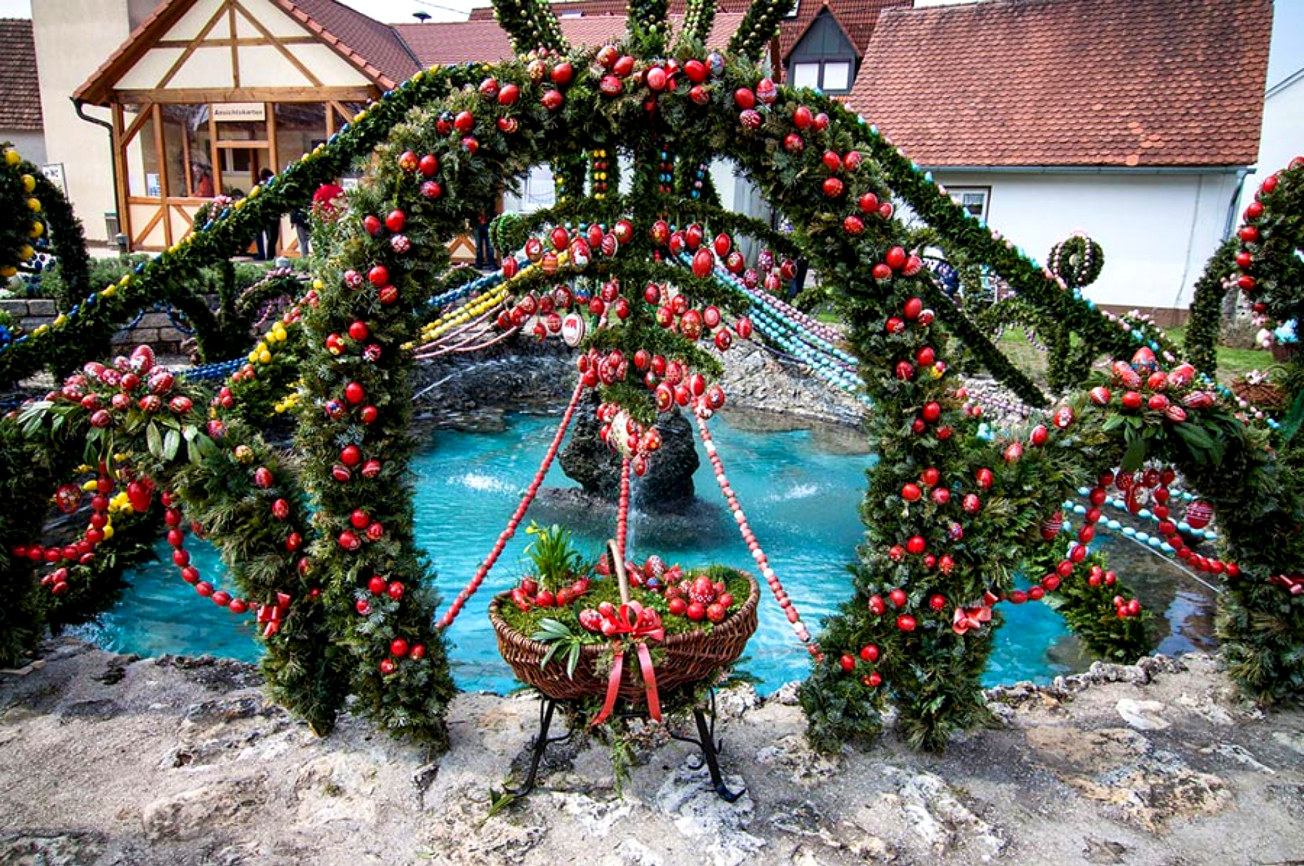 Stunning Bavarian Osterbrunnen Easter fountain tradition in a German village town square featuring hundreds of hand-painted blue and white Easter eggs strung on evergreen garlands and arches with spring flowers, bronze statue, and historic church clock tower in the background — how Germany celebrates Easter with unique cultural customs and folk art