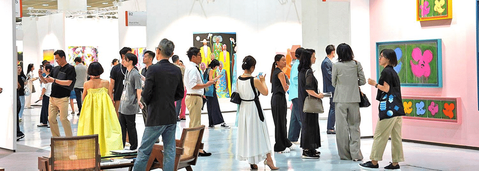Visitors gathered around contemporary artworks at an art exhibition, viewing paintings and installations in a bright gallery space.