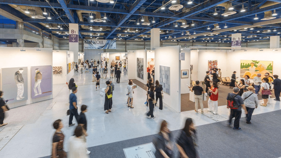 Busy contemporary art fair with multiple gallery booths, artworks on display, and visitors walking through the exhibition space.