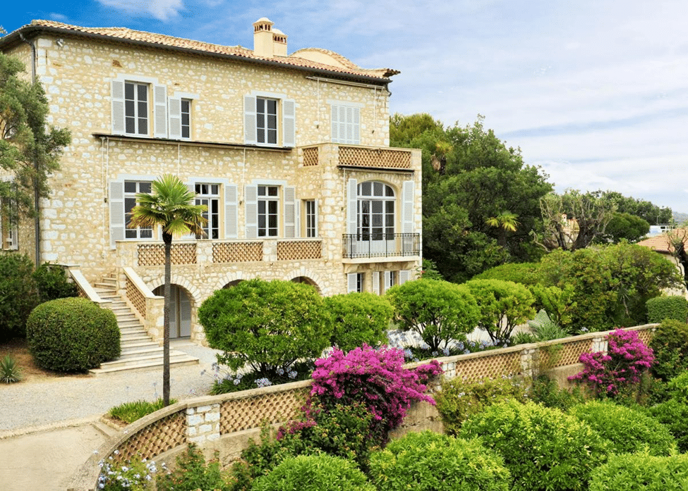 Large historic stone villa with shuttered windows, balconies, landscaped gardens, and flowering bushes under a blue sky.