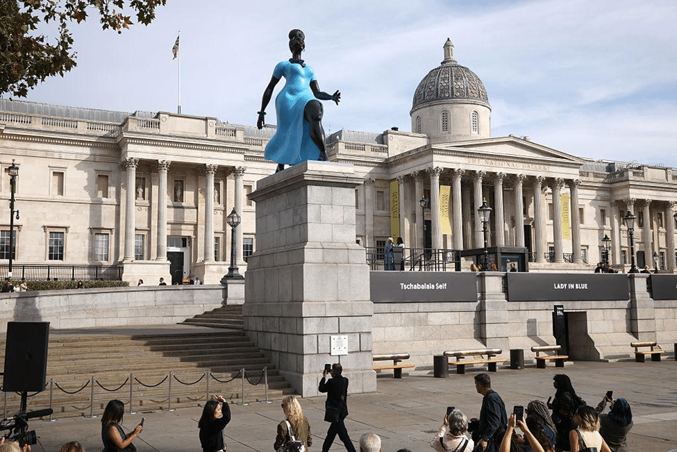 Blue painted-bronze sculpture standing on a plinth in front of the National Gallery in London, with visitors gathered in Trafalgar Square.