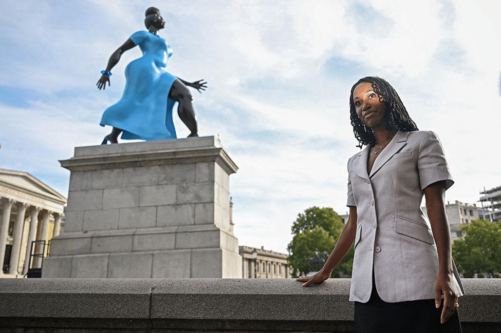 Woman standing beside the blue painted-bronze sculpture in Trafalgar Square, with the National Gallery visible in the background.