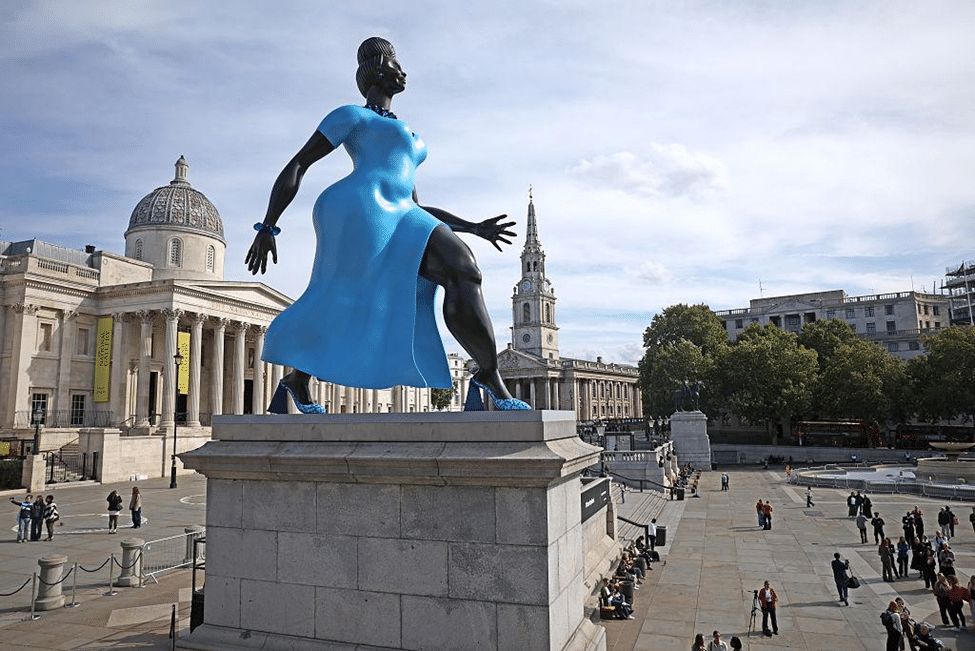 Blue painted-bronze sculpture viewed from behind on a stone plinth in Trafalgar Square, with the National Gallery and visitors in the background.