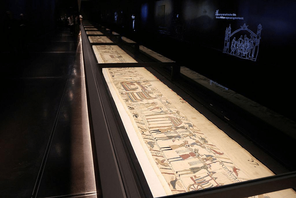 The Bayeux Tapestry displayed in a dark museum gallery, illuminated inside a long glass display case.