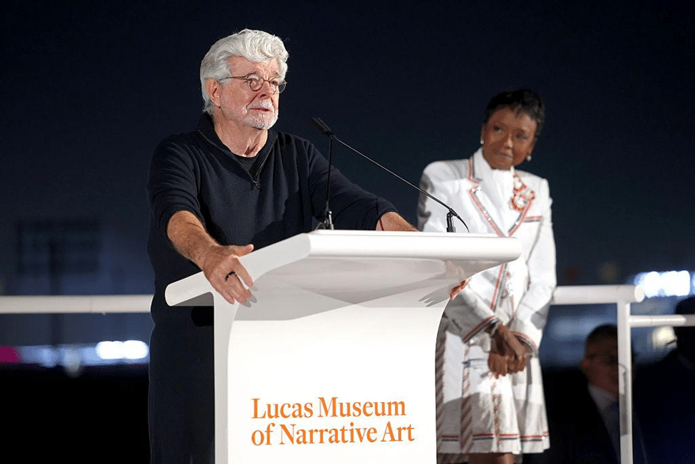 George Lucas speaking at a podium during the opening of the Lucas Museum of Narrative Art, with a guest standing behind him.