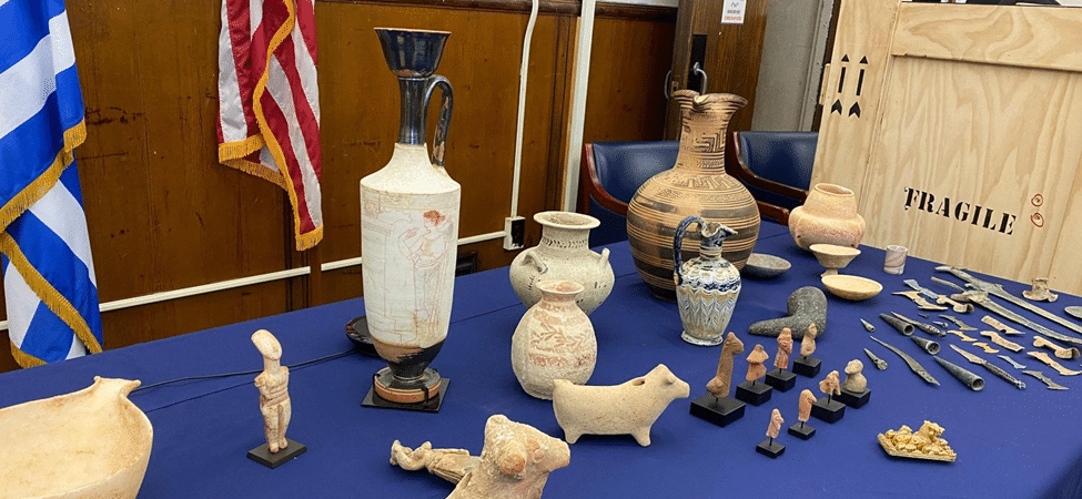 Museum display of African and ancient artifacts, including ceramic vessels, sculptural figures, and a large decorated vase arranged on a blue-covered table.