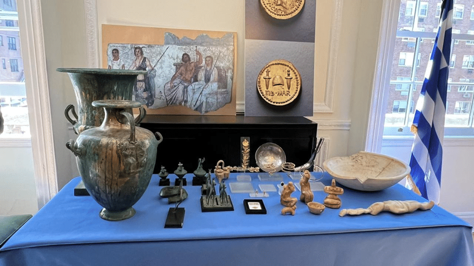 Museum display featuring paintings, carved plaques, sculptures, ceramic objects, and other historical artifacts arranged in cabinets and on a blue-covered table.