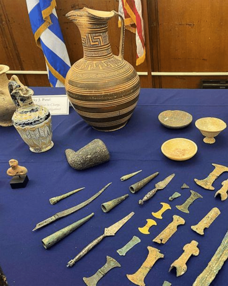 Museum display of ancient artifacts including a large decorated ceramic vessel, small pottery pieces, stone objects, and rows of metal tools and weapons on a blue-covered table.