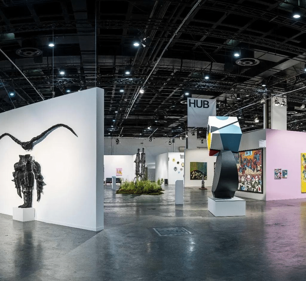 Interior view of an art fair with contemporary sculptures, paintings, and gallery booths.