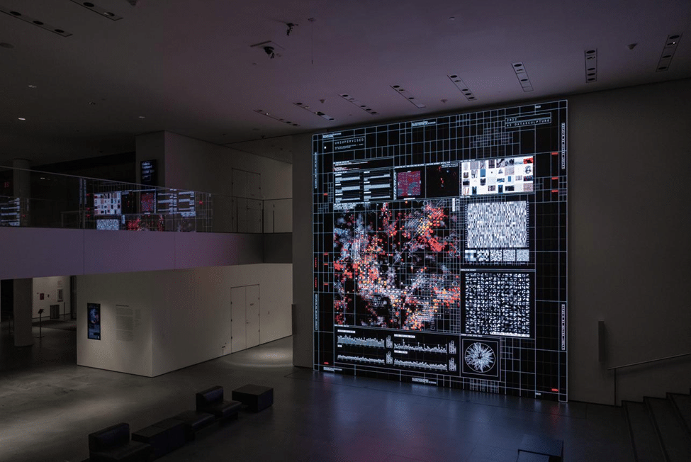 Visitors viewing a large digital artwork composed of intricate images and patterns on a screen in a dark gallery space.