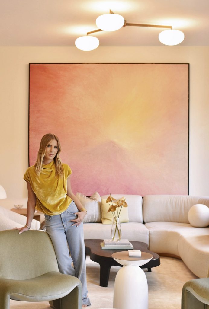 A woman in a mustard-yellow top and light blue jeans stands in a modern living room with a large pink abstract artwork behind her.