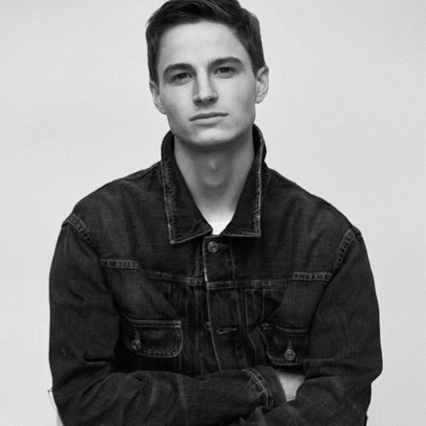Black-and-white portrait of a young man wearing a dark denim jacket, facing the camera with his arms crossed.