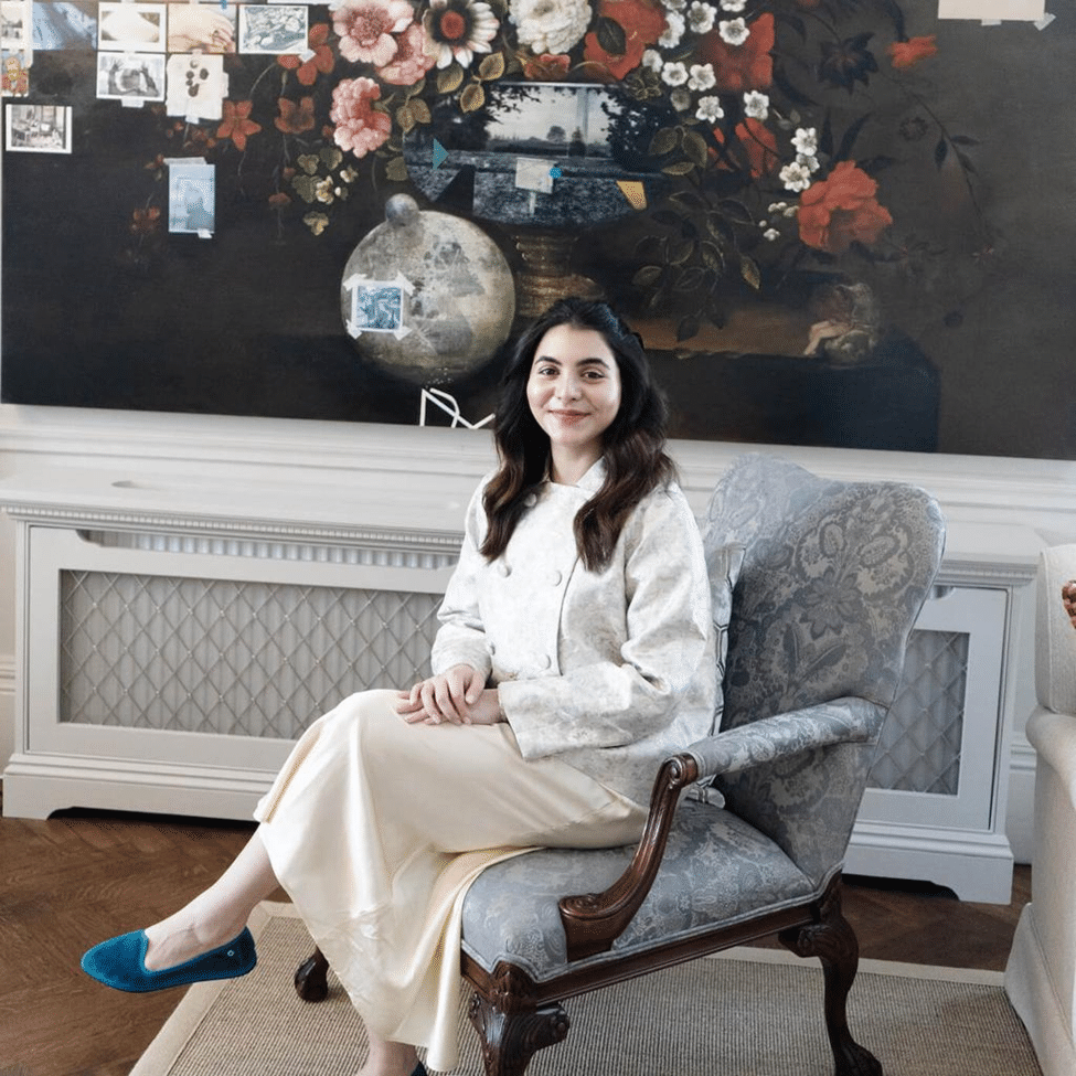 A woman in a white outfit sits in an armchair in front of a floral artwork and ornate interior decor.