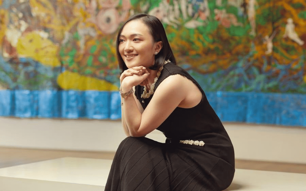 A woman in a black dress smiles while seated in front of a colorful abstract artwork.