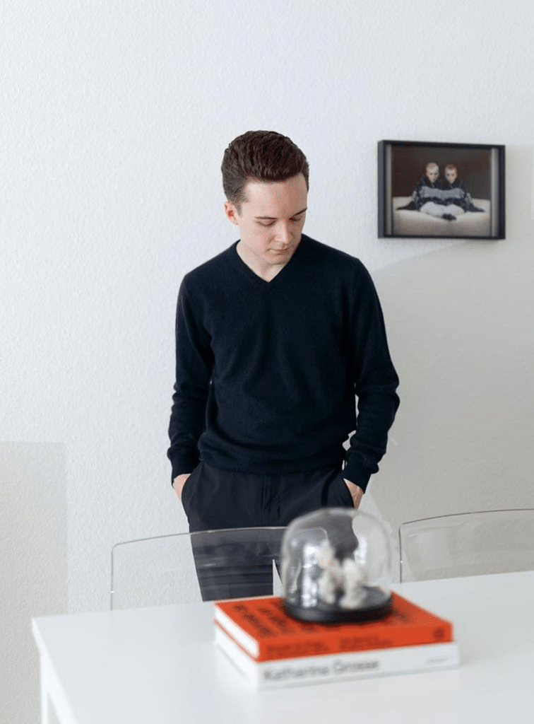 A young man in a dark sweater stands behind a table, looking down, with framed artwork on the wall behind him.