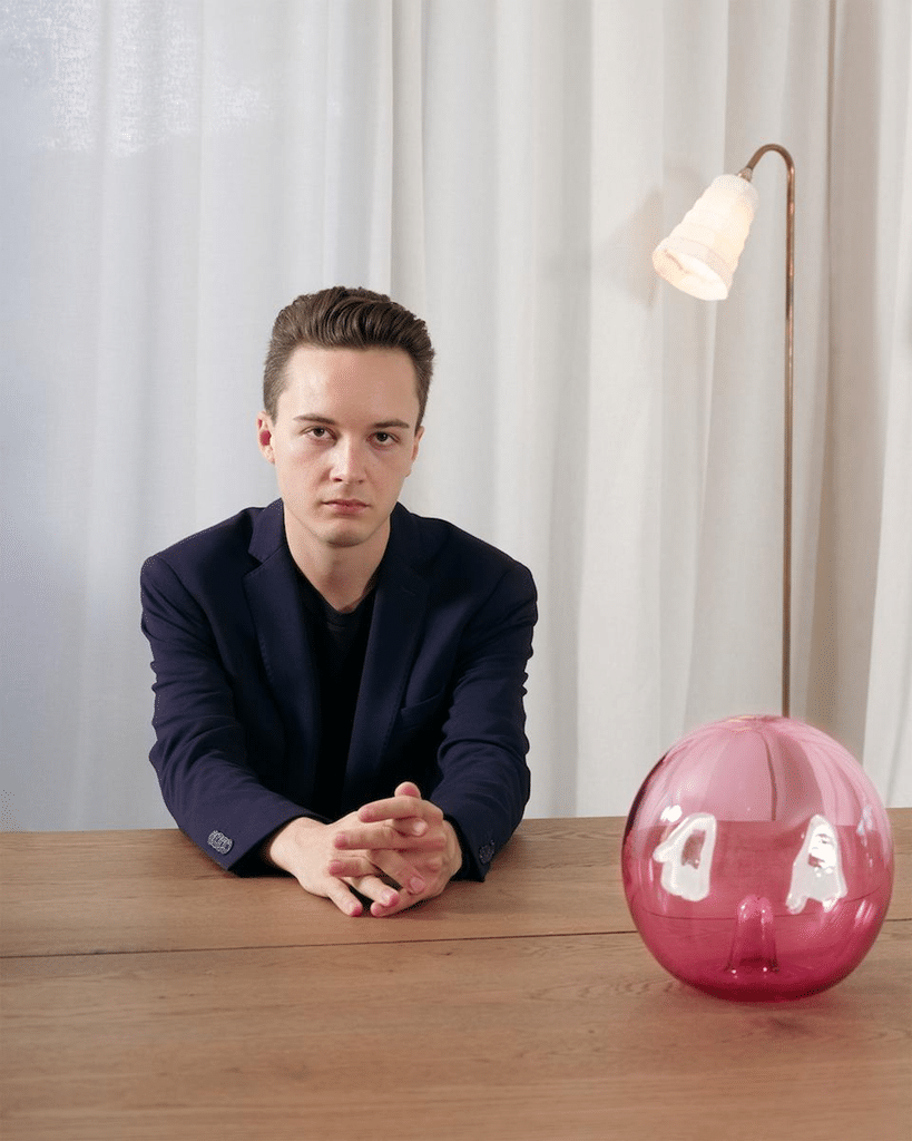 A young man in a dark blazer sits at a wooden table, facing the camera, with a pink glass vase and floor lamp beside him.