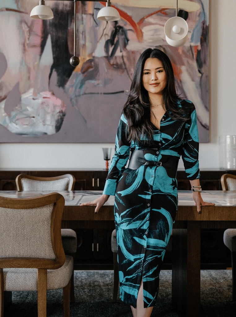 A woman in a patterned teal and black outfit stands in a modern dining area with a large abstract artwork behind her.