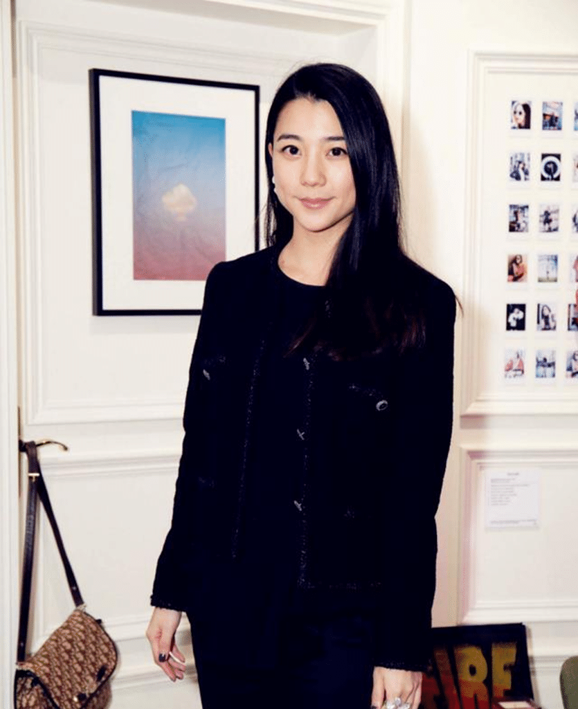 A woman in a black outfit stands in an elegant interior with framed artwork and photographs displayed on the walls.