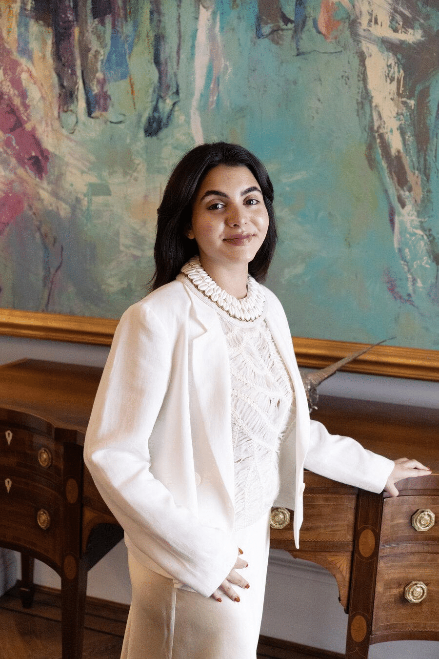 A woman in a white outfit stands in front of a large abstract painting in an elegant interior.