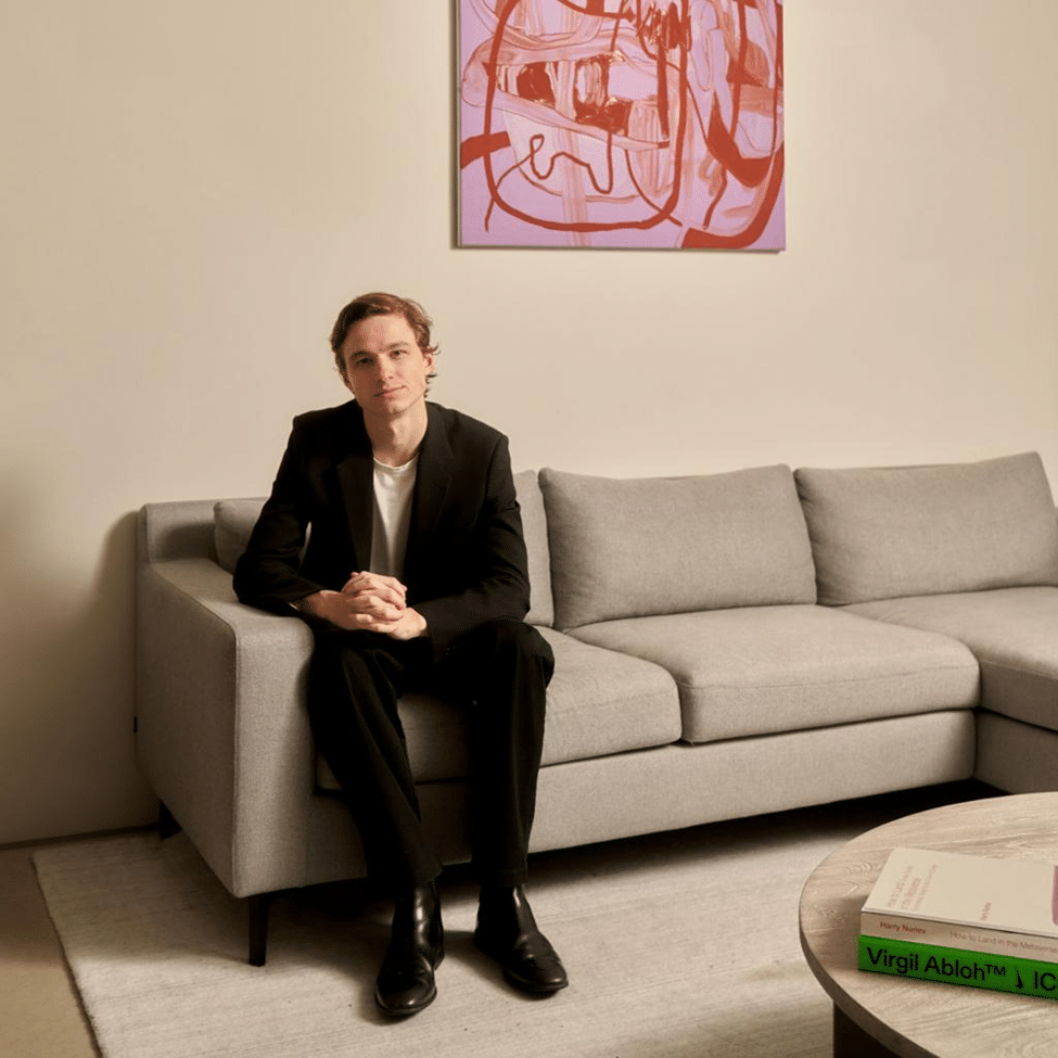 A man dressed in a black suit sitting on a beige sofa beneath a framed abstract artwork in a modern living room.
